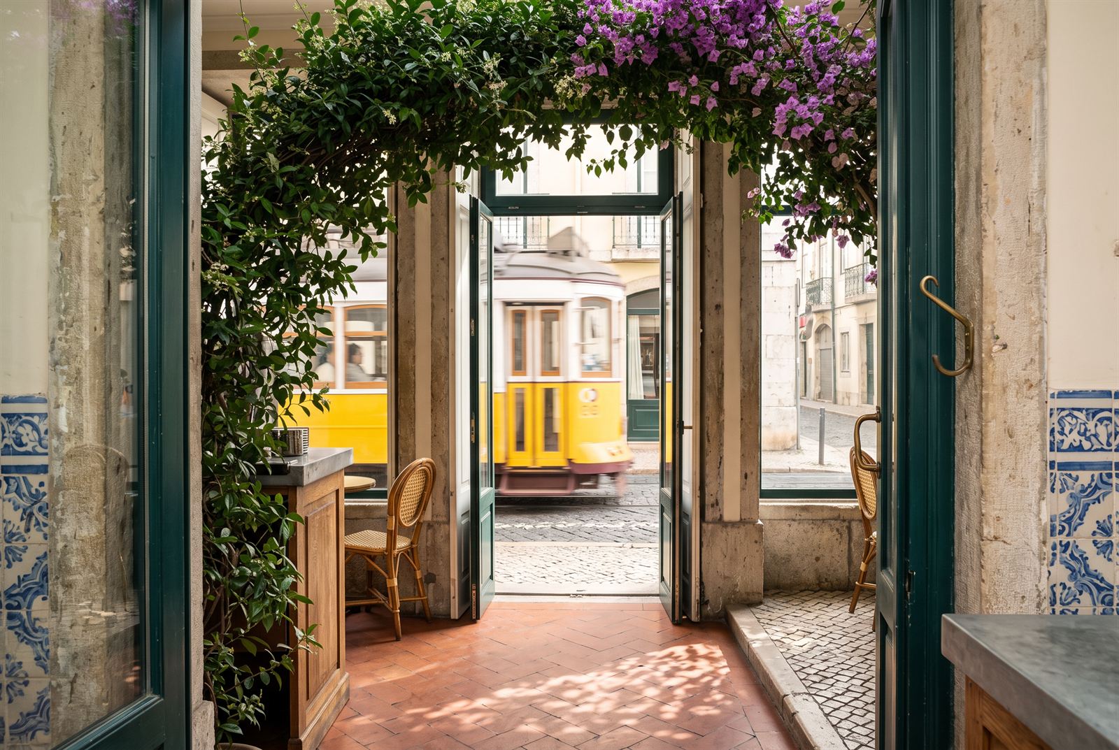View from inside the café through jasmine and bougainvillea over the door as a yellow Lisbon tram passes on the cobbles