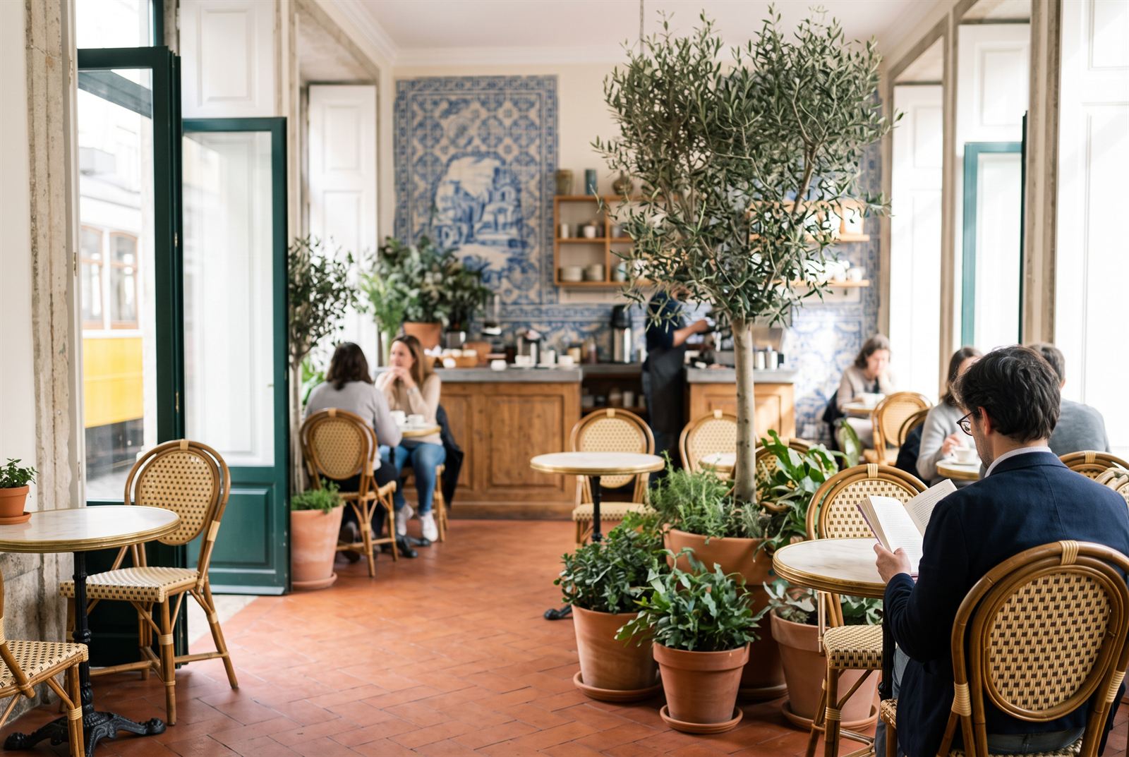 Café floor mid-morning — guests at marble bistro tables among cane chairs, potted olive tree and an azulejo mural behind the counter