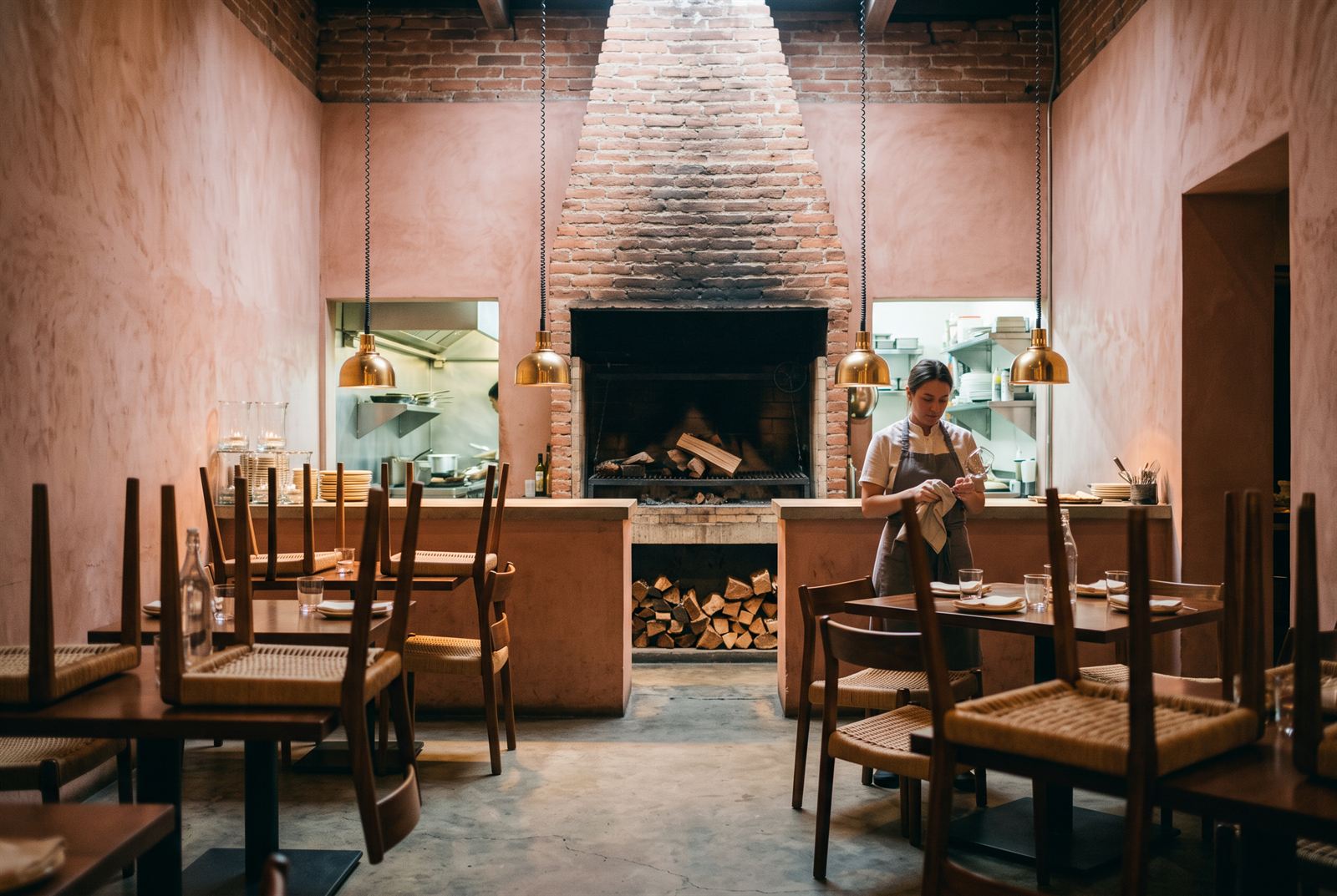 Morning prep — chairs still up on tables, a waiter polishing glasses, firewood stacked ready beneath the hearth